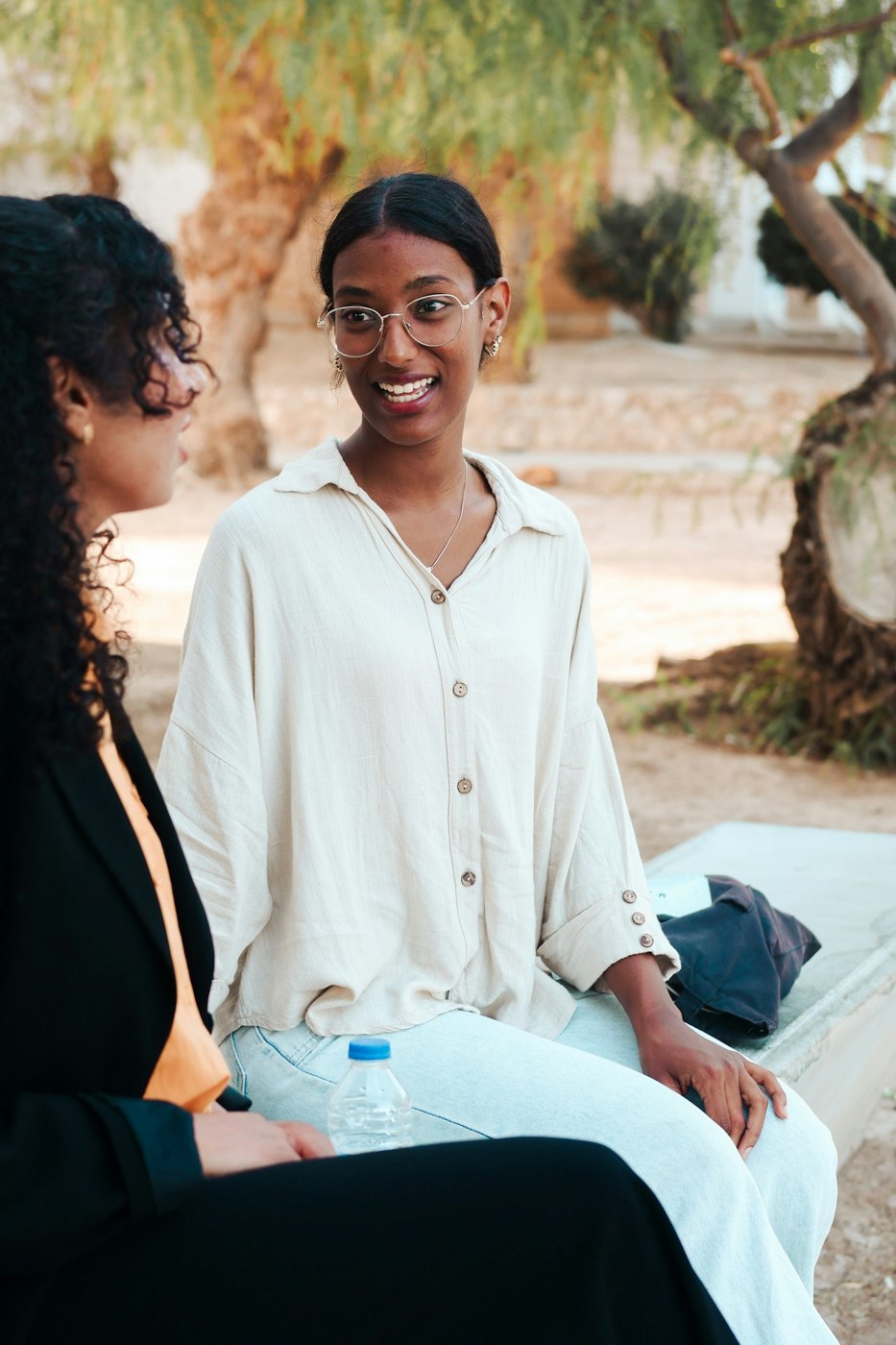 Two women smiling and talking on a bench beneath trees